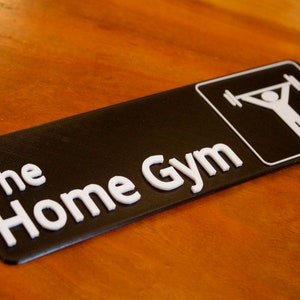 May include: Black rectangular sign with the words "The Home Gym" in white, 3D-style lettering. A white graphic of a person lifting weights is in the upper right corner. The sign is on a wooden surface.