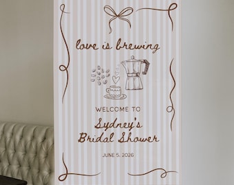 Può includere: Un cartello verticale su un cavalletto di legno con la scritta "love is brewing" e "Welcome to Sydney's Bridal Shower, June 5, 2026". Il cartello ha uno sfondo a righe e illustrazioni a tema caffè.