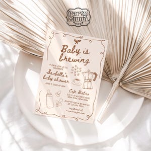 Puede incluir: Una invitaci&oacute;n de baby shower marr&oacute;n y blanca con tem&aacute;tica de caf&eacute;. La invitaci&oacute;n dice "Baby is brewing" e incluye una cafetera, granos de caf&eacute; y un biber&oacute;n. La invitaci&oacute;n es para el baby shower de Isabella el 5 de junio de 2026 a las 2 PM. La invitaci&oacute;n tambi&eacute;n incluye la ubicaci&oacute;n, Cafe Bistro, y la informaci&oacute;n de RSVP.