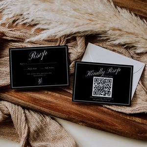 May include: Two black RSVP cards with white text and a QR code, resting on a wooden tray with a beige cloth and dried plants. One card says "Kindly RSVP" and the other says "RSVP".