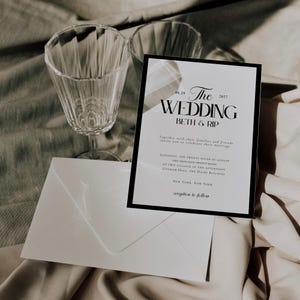 May include: A wedding invitation with the text "The Wedding" and the names "Beth & Rip" in black font. The invitation is in a black frame, with a white envelope and two clear wine glasses in the background. The invitation is on a light-colored fabric.