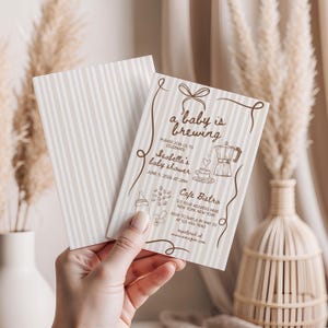 May include: Baby shower invitation with the text "a baby is brewing" and "Isabella's baby shower". The invitation has a striped background and features illustrations of a coffee pot, coffee cup, and baby bottle. The invitation is held in a person's hand.