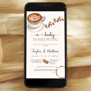 Pode incluir: Um smartphone branco com um convite para um chá de bebê na tela. O convite apresenta uma xícara de café com um coração e o texto "a baby is brewing". O convite também inclui os nomes "Taylor & Nathan" e a data "sábado, 25 de outubro, 14:00".
