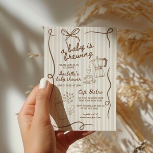 May include: A baby shower invitation card with a beige and white striped background. The card features brown decorative elements, including a bow, swirls, and illustrations of a coffee pot, coffee beans, and a baby bottle. Text on the card reads "a baby is brewing".