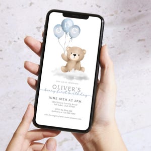 May include: A smartphone screen shows a first birthday invitation. The design includes a watercolour teddy bear holding blue balloons spelling "one." The invitation reads "Oliver's beary first birthday" with event details, including the date, time, and location.
