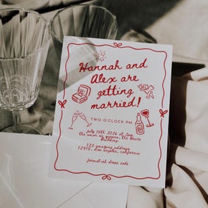 May include: A white wedding invitation with red text and illustrations. The text reads "Hannah and Alex are getting married! Two O'Clock PM July 15th 2026 at 2pm the amor on queens, the Haire Building. 123 gmazing address 12345, Los angeles, california formal-ish dress code"