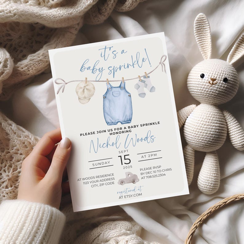 May include: A baby sprinkle invitation with a blue and white color scheme. The invitation features a clothesline with a baby's outfit hanging on it. The text on the invitation reads "It's a baby sprinkle!" and "Please join us for a baby sprinkle honoring Nichol Woods".