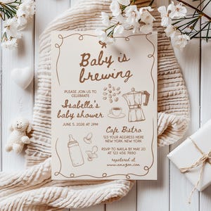 Puede incluir: Una invitaci&oacute;n de baby shower de color marr&oacute;n y blanco con una ilustraci&oacute;n de una taza de caf&eacute; y una cafetera. El texto dice "Baby is brewing" e incluye los detalles del evento, incluyendo la fecha, la hora, la ubicaci&oacute;n y la informaci&oacute;n de RSVP. La invitaci&oacute;n tambi&eacute;n incluye el texto "registered at www.amazon.com".