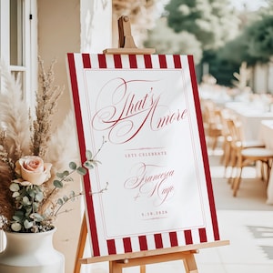 Op de afbeelding: Een welkomstbord voor een bruiloft met een rood-witte gestreepte rand en de woorden "That's Amore" in sierlijk schrift. Het bord bevat ook de namen Francesca en Hugo, en de datum 5.10.2026.