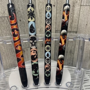 May include: Four black pens with different Magic: The Gathering symbols on them. The symbols are a sun, a drop of water, a skull, and a tree. The pens are on a clear plastic stand.