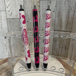 May include: Three pens with pink and white designs, displayed in a clear acrylic pen holder. One pen features a pink heart and the word "Barbie", another has a pink and black pattern, and the third has the word "Barbie" repeated.