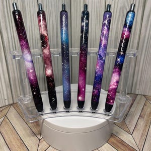 Puede incluir: Un juego de seis bolígrafos con temática de galaxia con acabado plateado. Los bolígrafos están dispuestos en un soporte de plástico transparente sobre una base circular blanca. Los bolígrafos presentan una variedad de diseños de galaxias con tonos de rosa, morado y azul.