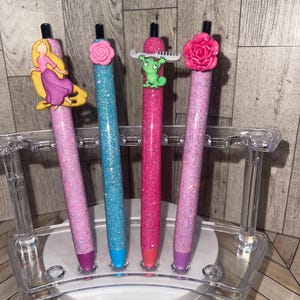 May include: Four glitter pens in a clear acrylic holder. The pens are pink, blue, and red, each with a different decorative topper: a princess, a rose, a frog, and another rose. The pen tips are colored to match the pen bodies.