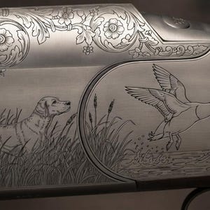 May include: A shotgun receiver featuring intricate silver-toned metal engravings. The design displays a hunting dog standing in tall grass alongside a flying duck over water, surrounded by detailed floral scrollwork and dark wooden stock.