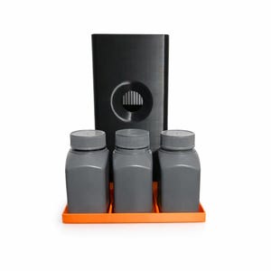 Op de afbeelding: Een zwarte dispenser met een cirkelvormige opening en drie grijze flessen met grijze deksels, geplaatst op een oranje dienblad. De dispenser is rechthoekig en staat achter de flessen.