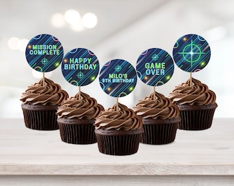 May include: Five chocolate cupcakes with neon green and blue themed cupcake toppers. The toppers have text that reads "Mission Complete", "Happy Birthday", "Milo's 9th Birthday", "Game Over", and a target with a star in the center.