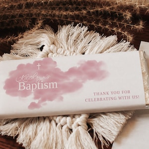 Może przedstawiać: Białe opakowanie czekolady z napisem "Kathryn's Baptism" w ozdobnej czcionce, różowym wzorem chmury i krzyżem. Na opakowaniu znajduje się również napis "Dziękujemy za świętowanie z nami!"