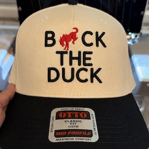 May include: A cream-colored baseball cap with a black brim featuring the embroidered black text "BACK THE DUCK" with a red bucking horse icon replacing the letter "U". A rectangular sticker on the brim displays the brand name OTTO and product details.
