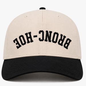 May include: A cream-colored baseball cap with a black brim and black embroidered text that reads BRONC-HOE in an arched, upside-down orientation across the front panels. The cap features a structured crown and a top button.
