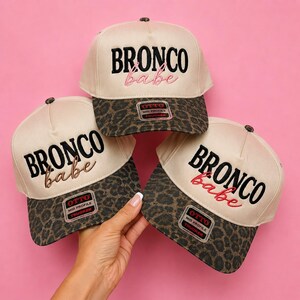 May include: Three beige trucker hats with leopard print brims. Each hat features the embroidered text "BRONCO" in black block letters and "babe" in cursive script. The hats include a rectangular sticker on the brim reading "OTTO MID PROFILE TRUCKER".