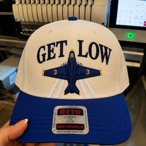 May include: A white baseball cap with a blue brim featuring an embroidered blue jet aircraft in the center with the number 3 on its wings. The text GET LOW is embroidered in blue above the jet. A silver sticker on the brim reads OTTO.