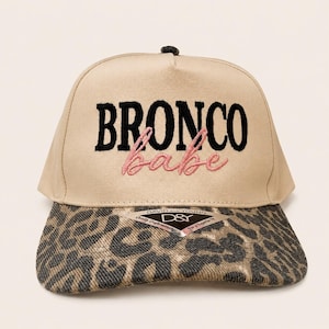 May include: A beige baseball cap featuring the embroidered text BRONCO in black and babe in pink. The brim displays a brown and black leopard print pattern. A triangular sticker with the text DAVID AND YOUNG D&Y Quality Craftsmanship is attached.