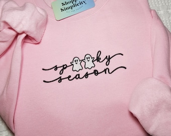 May include: A light pink sweatshirt with the words "spooky season" in black script, featuring two small white ghost illustrations. The sweatshirt has long sleeves and a crew neck. A small tag with the text "Shoppe Simplicity" is attached.