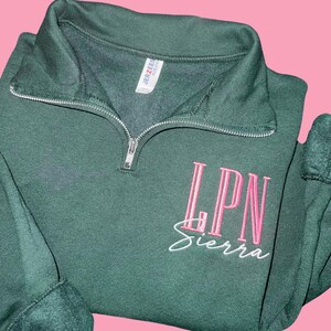 May include: A dark green quarter-zip sweatshirt with the embroidered letters "LPN" in pink, above the name "Sierra" in white script. The sweatshirt has a ribbed collar and cuffs, and a silver zipper.