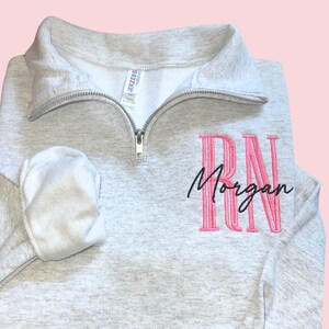 Puede incluir: Una sudadera gris claro con cuello con cremallera. La sudadera presenta las letras bordadas "RN" en rosa, con el nombre "Morgan" bordado debajo en escritura negra. Las mangas est&aacute;n dobladas y la etiqueta de la marca es visible.