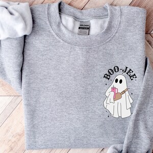 May include: A heather gray sweatshirt featuring a cartoon ghost with the text "Boo-jee". The ghost is holding a pink drink and wearing a brown fanny pack. The design includes small star accents.