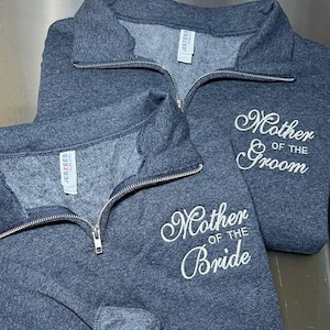May include: Two charcoal gray quarter-zip sweatshirts featuring white embroidered text. One sweatshirt displays the words Mother of the Groom, and the other displays the words Mother of the Bride. Both garments have silver-toned metal zippers.