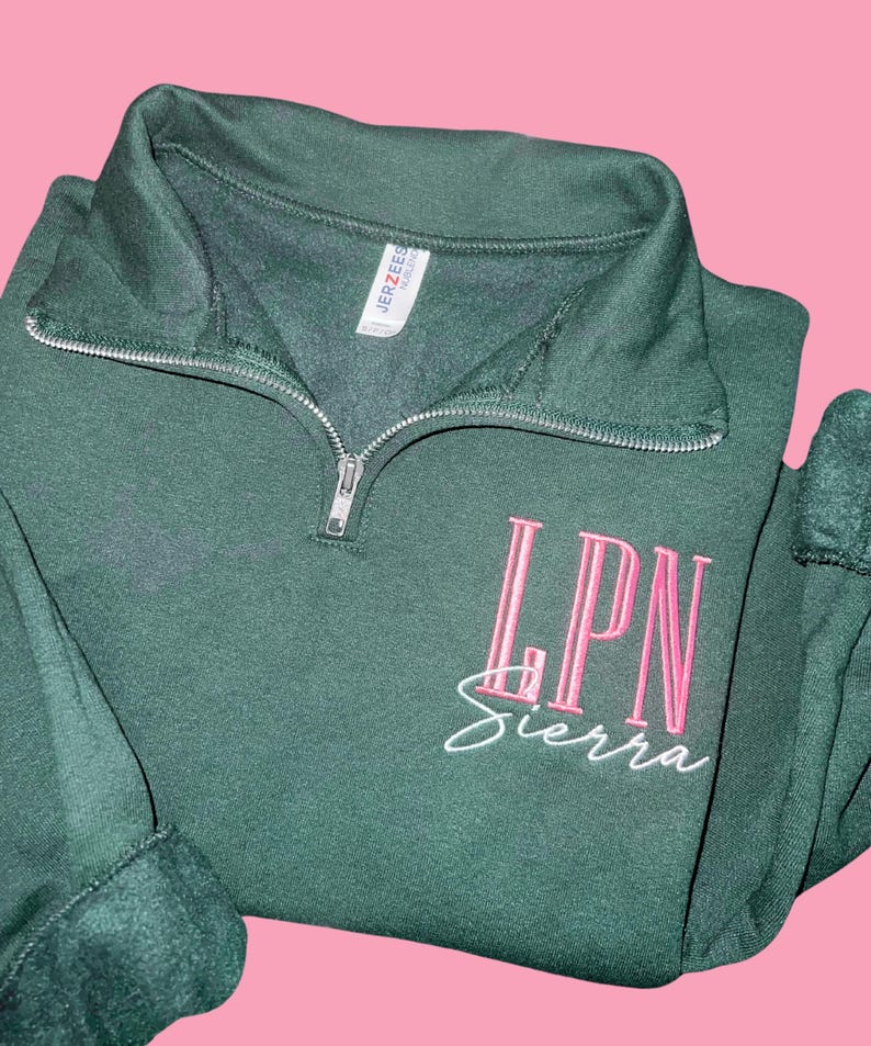 May include: A dark green quarter-zip sweatshirt with the embroidered letters "LPN" in pink, above the name "Sierra" in white script. The sweatshirt has a ribbed collar and cuffs, and a silver zipper.