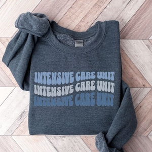 Peut inclure: Un sweat-shirt gris fonc&eacute; avec les mots "INTENSIVE CARE UNIT" r&eacute;p&eacute;t&eacute;s trois fois dans une police r&eacute;tro bleue et grise.