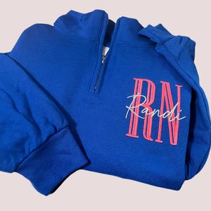 Puede incluir: Sudadera azul real con cremallera de un cuarto y mangas largas. La sudadera presenta el nombre "Randi" bordado en escritura blanca sobre un dise&ntilde;o "RN" rosa. La cremallera es negra.