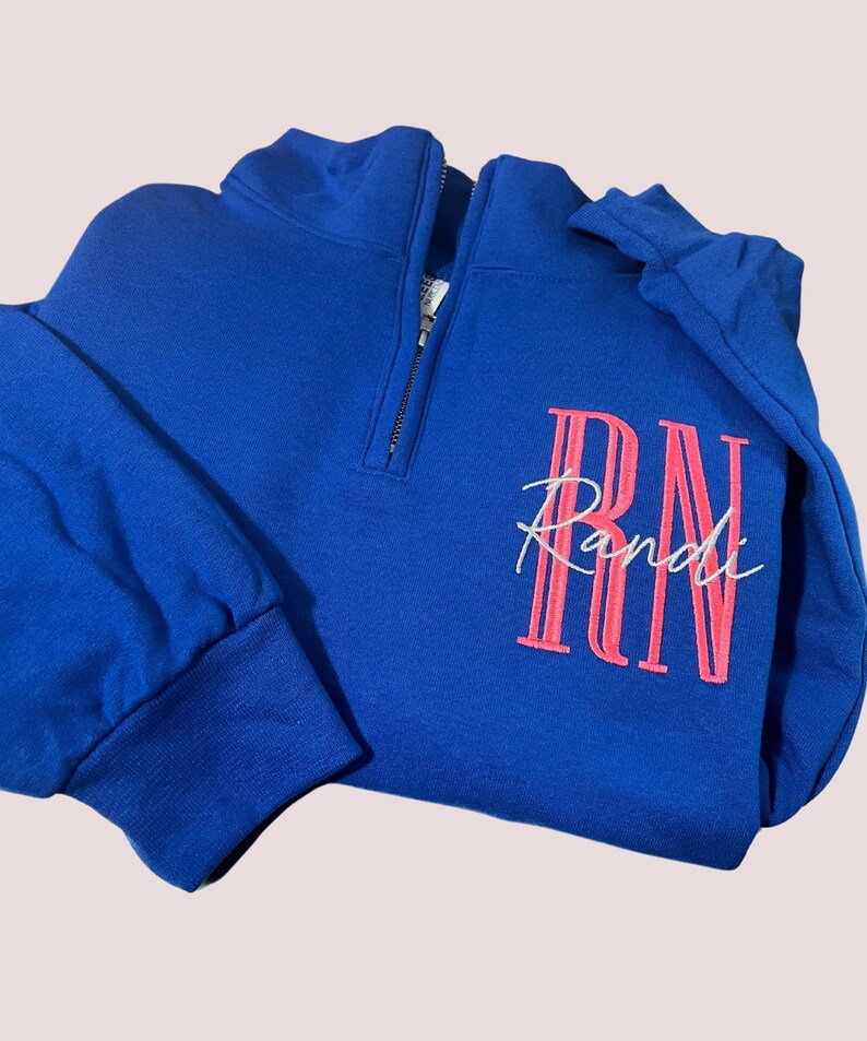 Puede incluir: Sudadera azul real con cremallera de un cuarto y mangas largas. La sudadera presenta el nombre "Randi" bordado en escritura blanca sobre un dise&ntilde;o "RN" rosa. La cremallera es negra.