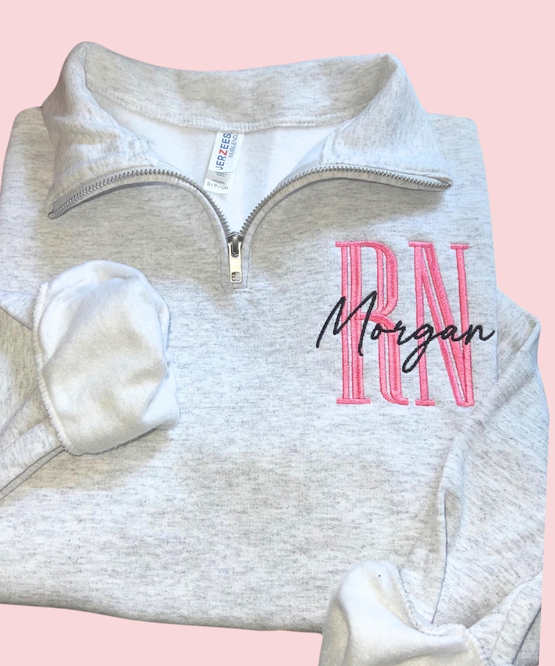 Puede incluir: Una sudadera gris claro con cuello con cremallera. La sudadera presenta las letras bordadas "RN" en rosa, con el nombre "Morgan" bordado debajo en escritura negra. Las mangas est&aacute;n dobladas y la etiqueta de la marca es visible.