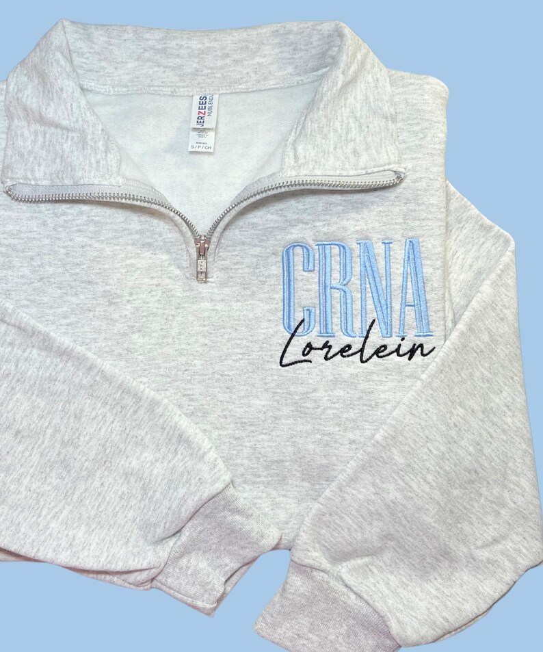 May include: A gray zippered sweatshirt with the text 'CRNA Lorelein' embroidered on the front. The text 'CRNA' is in blue and the text 'Lorelein' is in black.