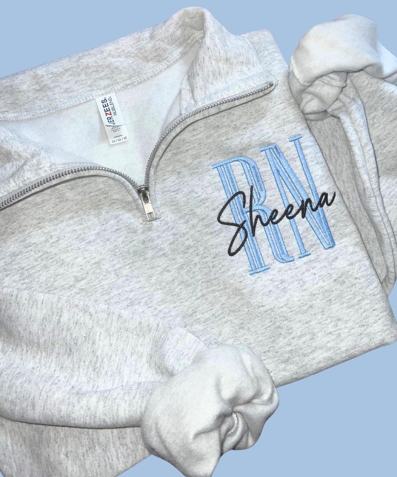 Puede incluir: Una sudadera gris claro con cremallera de un cuarto con el nombre "Sheena" bordado en escritura negra debajo de las letras azules "RN". La sudadera tiene un interior blanco y una cremallera plateada.