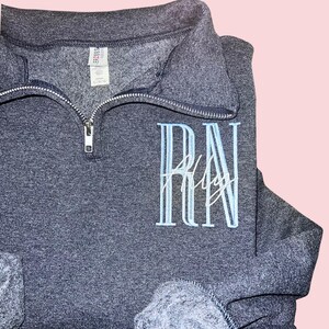 Puede incluir: Una sudadera gris oscuro con cuello con cremallera. La sudadera presenta las letras bordadas "RN" en azul claro, con el nombre "Ally" bordado debajo en escritura blanca. Las mangas est&aacute;n dobladas.