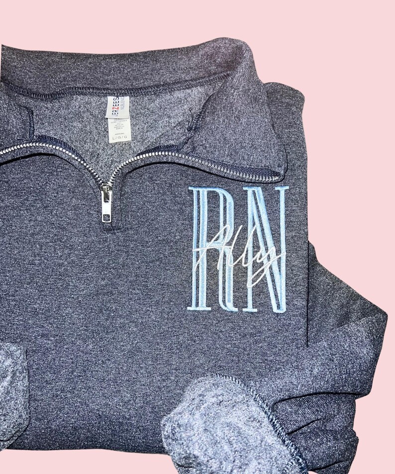 Puede incluir: Una sudadera gris oscuro con cuello con cremallera. La sudadera presenta las letras bordadas "RN" en azul claro, con el nombre "Ally" bordado debajo en escritura blanca. Las mangas est&aacute;n dobladas.