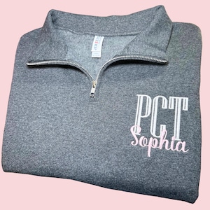 May include: A gray quarter-zip sweatshirt with the text "PCT Sophia" embroidered on the front in white and pink.
