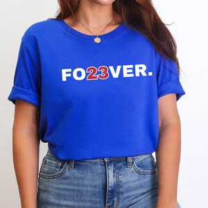 May include: Royal blue t-shirt with the text "FO23VER." in white and red. The shirt has short sleeves and is paired with blue jeans. The overall style is casual and comfortable.