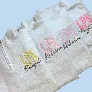 May include: Several light gray quarter-zip sweatshirts with embroidered text. Each sweatshirt has "LPN" in a different color, with a name below. The colors include yellow, pink, and purple. The background is a light blue.