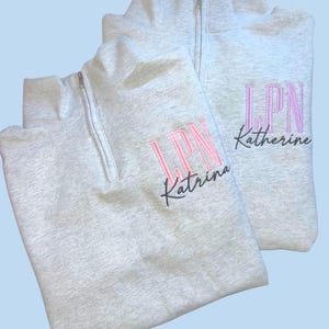 May include: Two light gray quarter-zip sweatshirts. Each sweatshirt has the embroidered letters "LPN" in pink, with a name below in a script font. One says "Katrina" and the other says "Katherine".