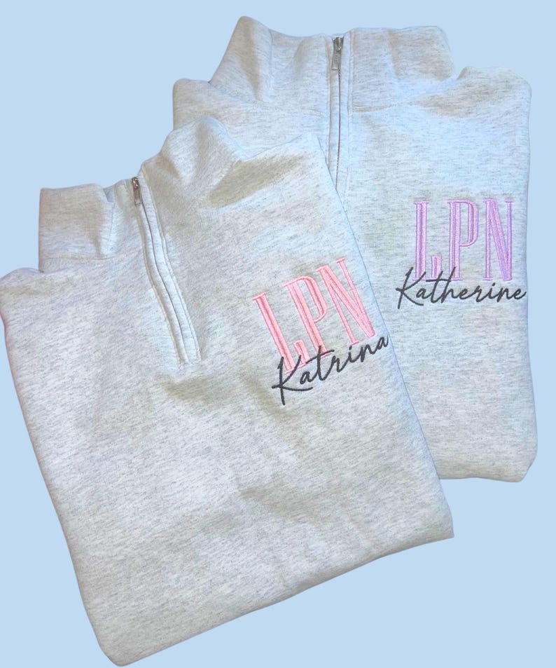 May include: Two light gray quarter-zip sweatshirts. Each sweatshirt has the embroidered letters "LPN" in pink, with a name below in a script font. One says "Katrina" and the other says "Katherine".