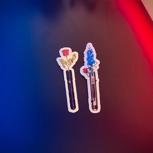 May include: Two stickers featuring a black lightsaber with a red rose and a blue flower on top. The stickers are on a blue and red background.