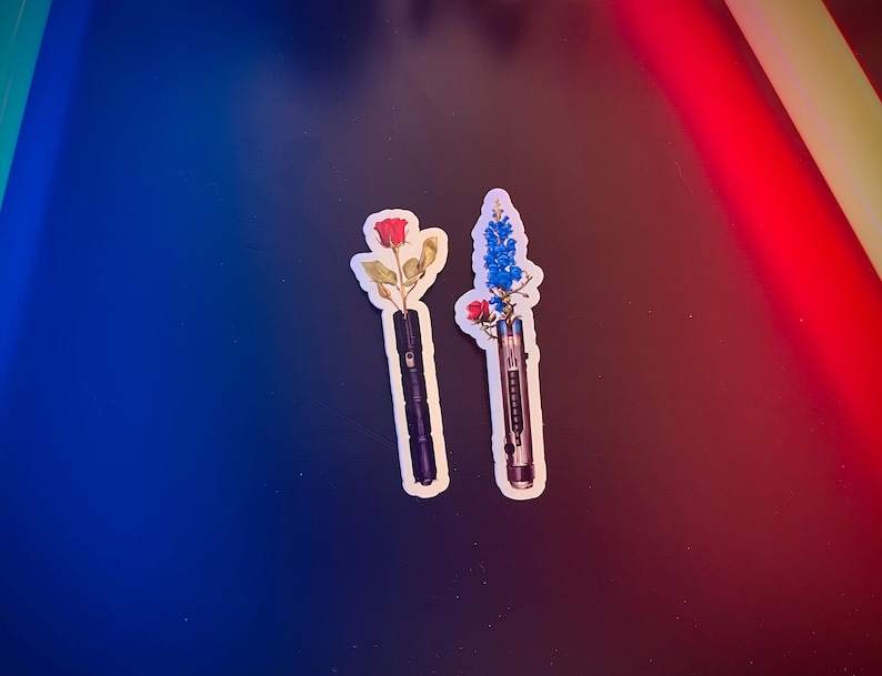 May include: Two stickers featuring a black lightsaber with a red rose and a blue flower on top. The stickers are on a blue and red background.