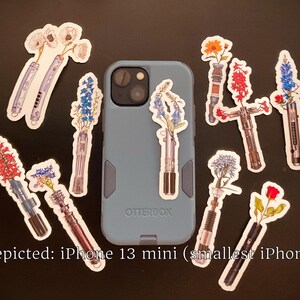 May include: A blue iPhone 13 mini phone case with a variety of floral stickers on it. The stickers feature different types of flowers in vases, including bluebells, roses, and lilies. The text "Otterbox" is visible on the case.