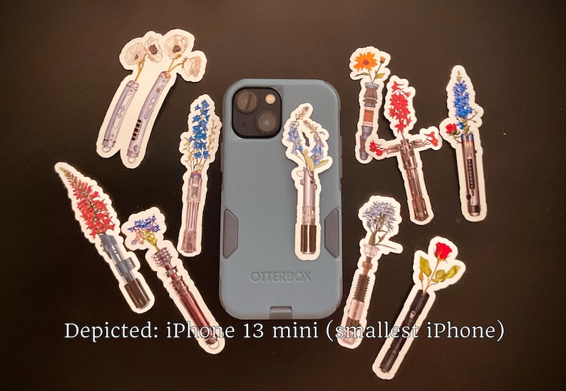 May include: A blue iPhone 13 mini phone case with a variety of floral stickers on it. The stickers feature different types of flowers in vases, including bluebells, roses, and lilies. The text "Otterbox" is visible on the case.