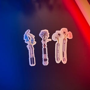 May include: Four stickers featuring different flowers in a lightsaber design. The flowers are blue, white, and pink. The lightsabers are silver and black.